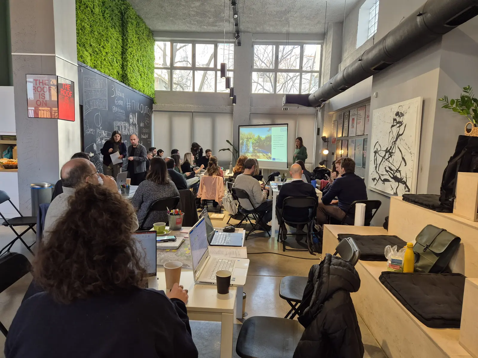 Workshop room with participants seated at tables, a presenter beside a projection screen and a planted green wall.