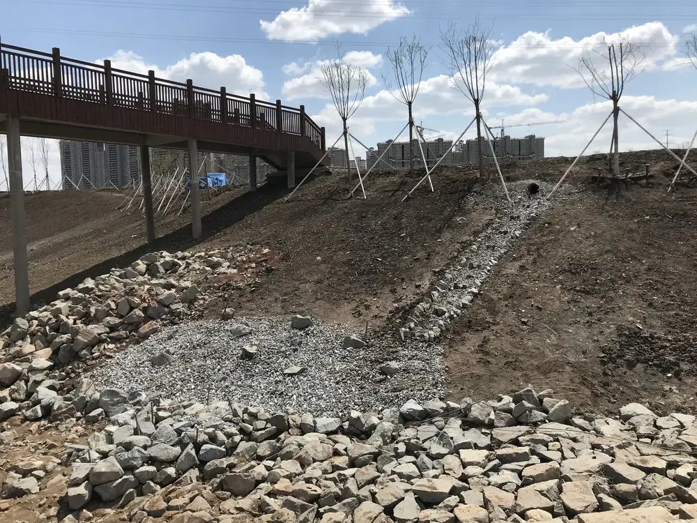 Holzsteg auf Betonstützen über einer frisch modellierten Böschung mit verankerten Jungbäumen und einer steingefassten Mulde.