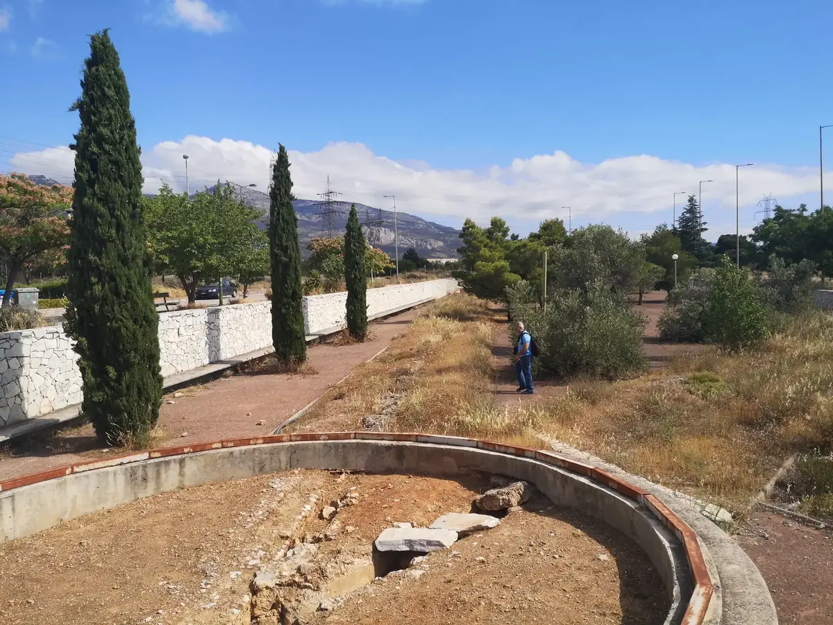 Dry circular basin beside a low stone wall, cypresses and olive trees along the aqueduct route, with mountains behind.
