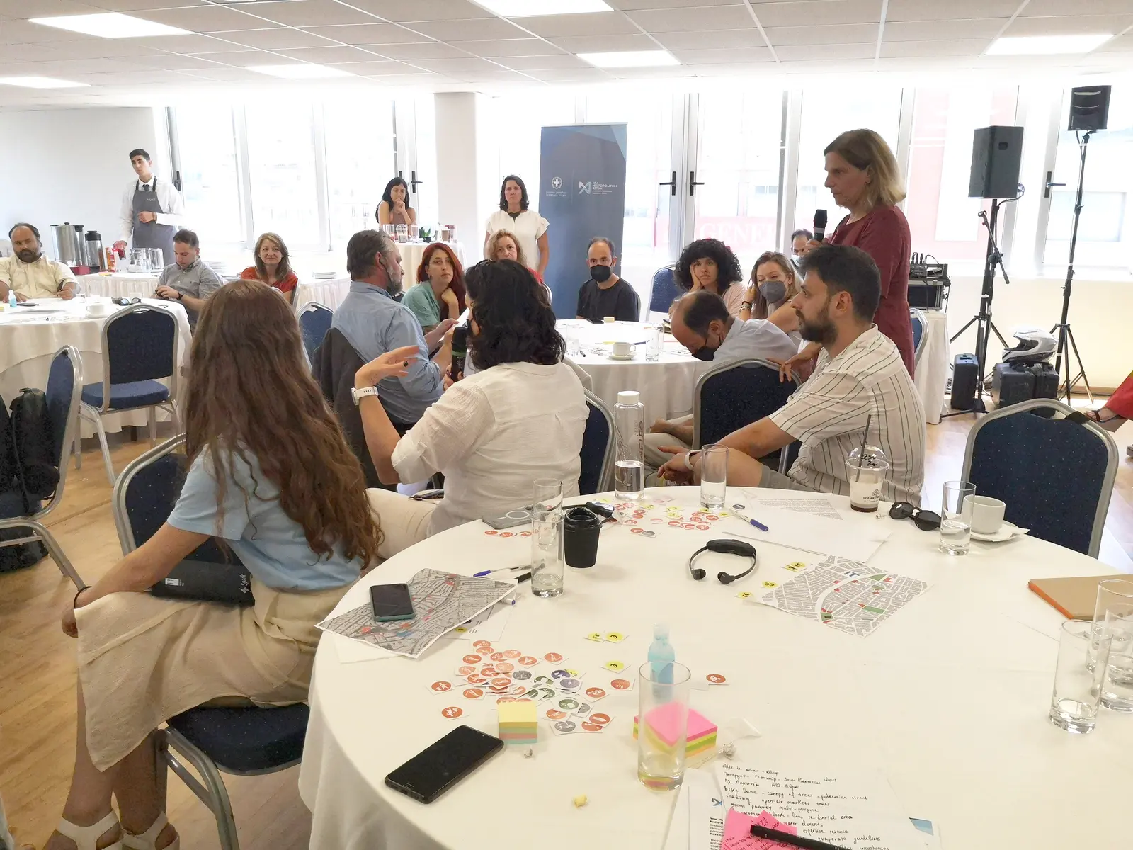Participants seated around round tables at a workshop, listening to a speaker with a microphone.