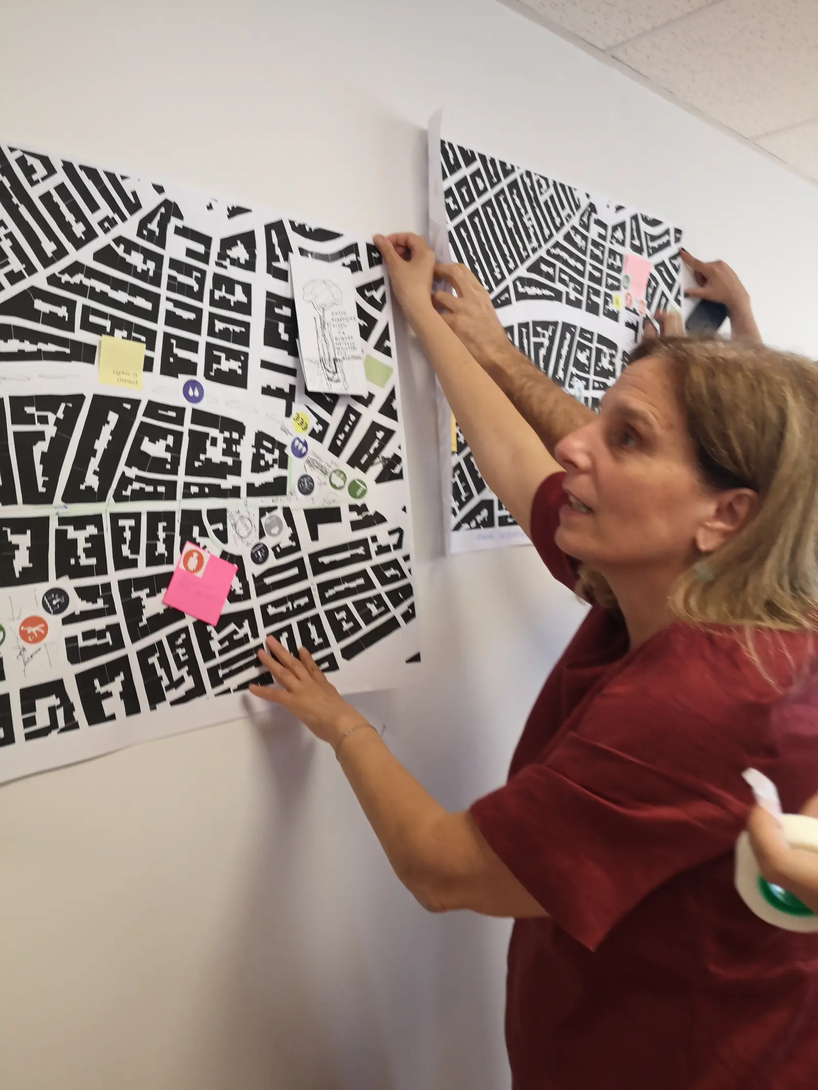 Two people pinning large black-and-white neighbourhood maps with stickers and notes to a wall.